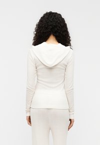 Λευκό μακρυμάνικο τοπ με κουκούλα, ριμπ ύφανση, εφαρμοστή γραμμή και ευθύ τελε ίωμα, σε συνδυασμό με παντελόνι που ταιριάζει.