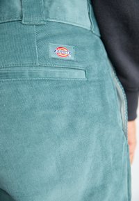 Zelene kordurojske nedrse s hrbtnim žepom in tkano oznako "Dickies". Tekstura je rebrasta z drobnimi črtami.