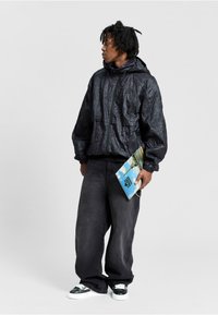 Schwarze Nylonjacke mit einer knitterigen Textur, ausgestattet mit einer Kapuze und Fronttaschen, kombiniert mit lockeren dunklen Hosen und kontrastierenden Sneakers.