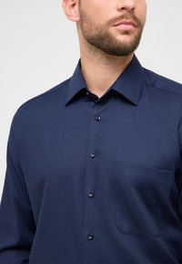 Camicia blu navy con bottoni, tessuto strutturato, maniche lunghe, colletto appuntito e taschino sul petto. Presenta bottoni scuri.