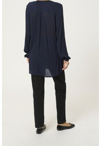 Marineblå langærmet top med afslappet pasform, der har en detalje med en søm bagpå, kombineret med sorte bukser og flade sorte sko. Glat stoftekstur.