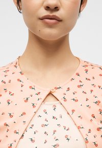 Κοντινό πλάνο ενός ατόμου που φοράει ένα ροδακινί φλοράλ πουκάμισο με κουμπιά, πάνω από ένα λευκό φλοράλ πουκάμισο, με ορατό λαιμό και γραμμή σαγονιού.