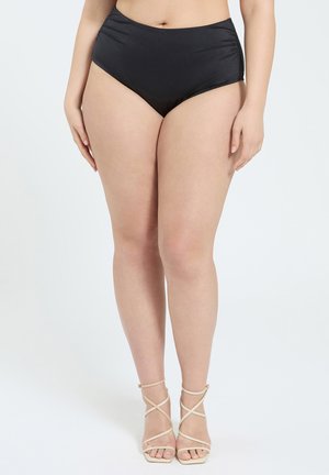 Nainen pukeutuneena korkeavyötäröisiin mustiin bikinin alaosiin ja beigeihin remmikkäisiin korkokenkiin seisoo valkoista taustaa vasten.