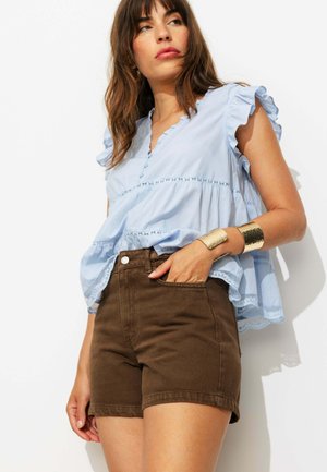 Femme portant un chemisier sans manches bleu clair à volants et un short marron taille haute, avec un bracelet manchette en or au poignet.