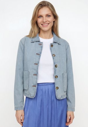 Lachende vrouw met een lichtblauwe spijkerjas over een wit shirt en een geplooide blauwe rok tegen een effen witte achtergrond.