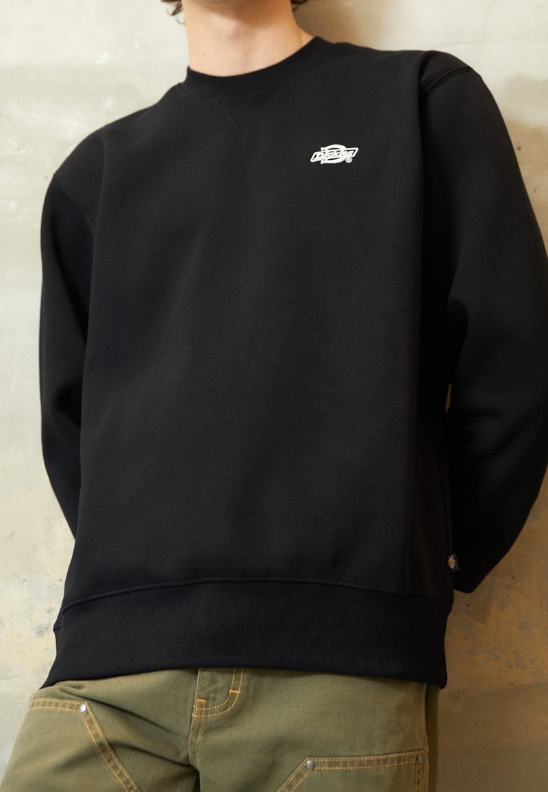 Personne portant un sweat-shirt noir avec un petit logo blanc sur la poitrine et un pantalon vert, debout contre un mur beige texturé.