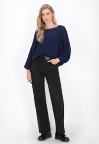 Frau mit langen blonden Haaren, die einen lockeren, navyfarbenen Strickpullover, schwarze hoch taillierte Jeans mit Gürtel und schwarze Stiefel trägt, steht vor einem weißen Hintergrund.