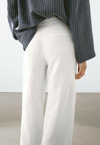 White pinstriped trousers made of a textured fabric, paired with a loose navy striped top. Features a back pocket and wide-leg design.