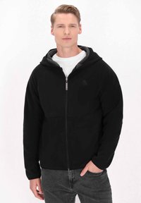 Svart zip-up hoodie med en gråfärgad luva, med en logotyp på bröstet, tillverkad av mjukt material med avslappnad passform och långa ärmar.