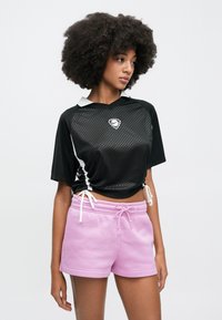 Μαύρο αθλητικό τοπ με υφασματικό σχέδιο, κοντά μανίκια και ρυθμιζόμενες πλευρές. Συνδυάζεται με ανοιχτό ροζ, άνετα σορτς με κορδόνι στη μέση.