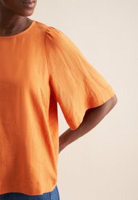 Top in lino arancione con maniche corte larghe e scollo rotondo. Presenta una texture morbida e una vestibilità rilassata, abbinato a pantaloni in denim.