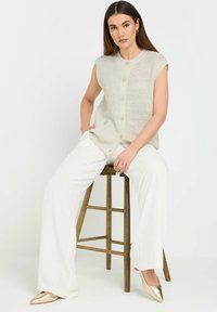 Gilet lavorato a maglia grigio chiaro con maniche corte e bottoni, indossato sopra pantaloni bianchi a gamba larga, abbinato a scarpe metalliche con punta a stiletto.