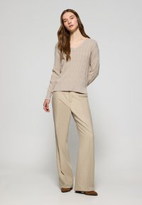 Beige kabelstickad tröja med V-ringning, kombinerad med hög midja beige byxor. Modellen står framför en neutral bakgrund och visar upp en avslappnad passform.