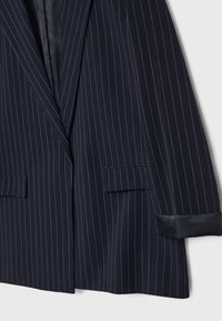 Donkerblauw pinstriped colbert met een gestructureerde vorm, voorzien van twee voorzakken en satijnen voering aan de manchetten. De stof heeft een zachte textuur.