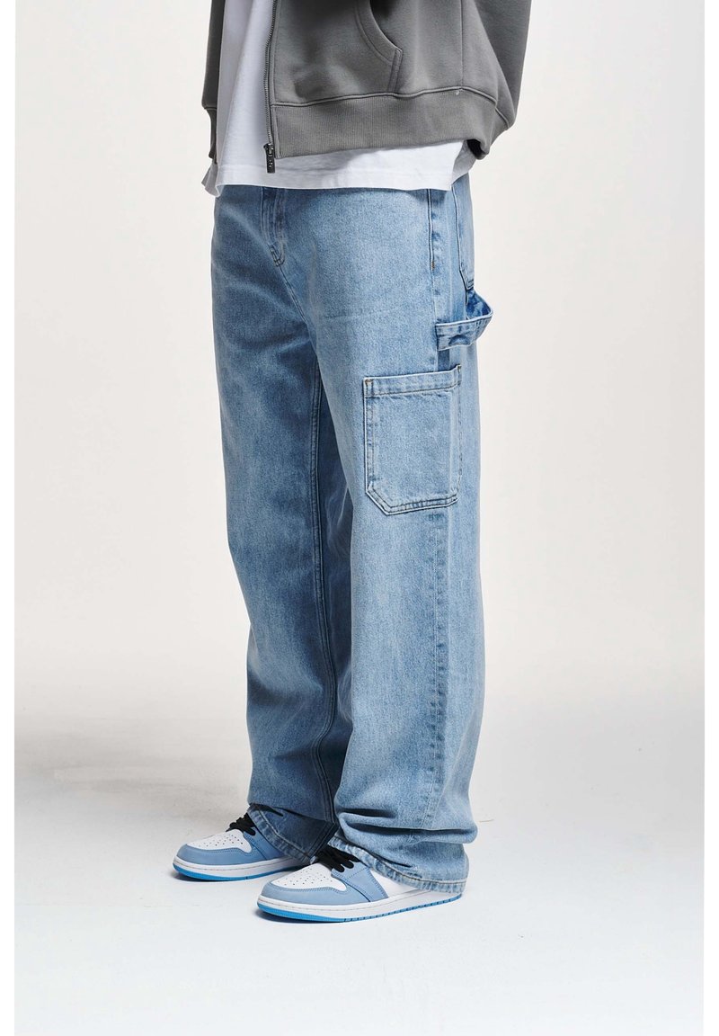 Pantalones cargo de mezclilla azul claro con un ajuste holgado, que cuentan con múltiples bolsillos y una textura desgastada. Combinados con zapatillas blancas y azules.