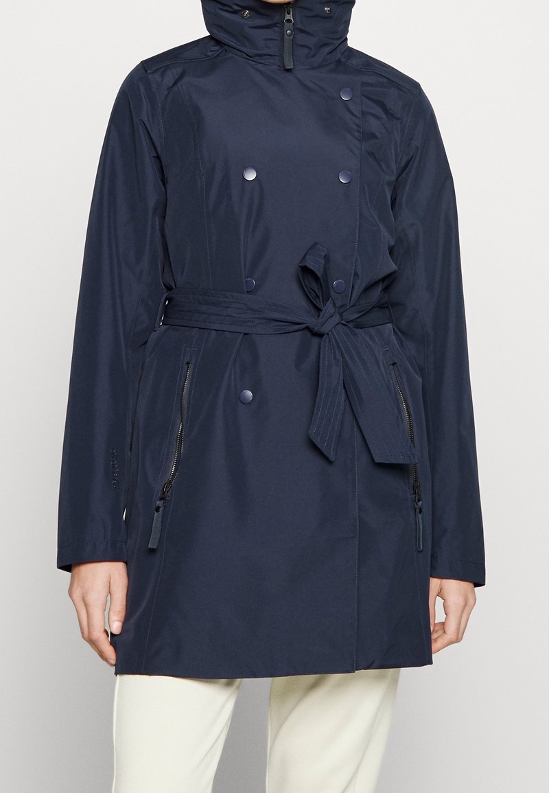 Imperméable bleu marine avec une coupe trench, doté d'une fermeture à double boutonnage, d'une ceinture réglable à la taille et de poches latérales zippées. Texture lisse.