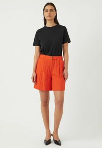 Musta lyhythihainen t-paita yhdistetty oransseihin laskostettuihin shortseihin, joissa on vyötärö ja etupainike. Mallilla on päällään mustat kärkitossut.