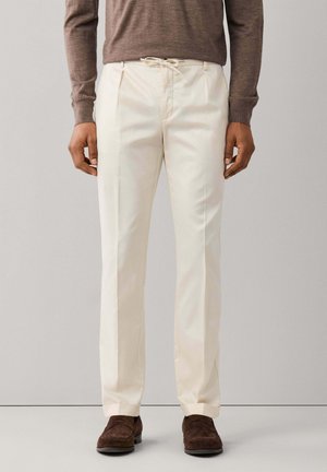 Homme portant un pantalon beige crème sur mesure avec taille à cordon, des mocassins en daim marron et un haut marron à manches longues.