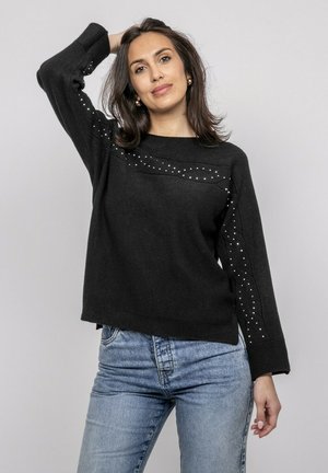 Femme portant un pull noir avec des détails cloutés et un jean bleu clair, posant avec un bras levé derrière la tête sur un fond gris.