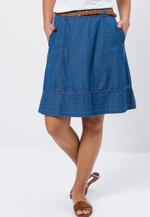 Donna con gonna di jeans blu al ginocchio, cintura intrecciata marrone e sandali bassi marroni, con le mani nelle tasche, in piedi con le gambe incrociate.