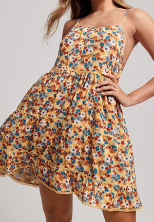 Mujer vestida con un vestido amarillo sin mangas con flores azules, naranjas y blancas, con falda con vuelos y un delicado borde en el bajo.