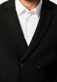 Blazer nero doppiopetto con rever a punta, realizzato in tessuto liscio. Presenta due bottoni neri e una camicia bianca con colletto sotto.