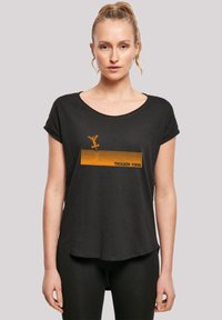 Svart t-skjorte med korte ermer laget av myk stoff, med et grafisk design i oransje som har teksten "TIGGER 1968" og et lekent designelement.