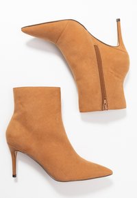 Paire de bottines en daim beige à bouts pointus avec talons stilettos, fermetures éclair latérales et texture lisse sur fond blanc.
