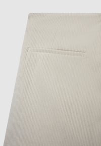 Tessuto di velluto a coste beige chiaro con coste verticali. Presenta una piccola tasca cucita sul retro. Texture morbida, design semplice.