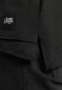 Svart bomull T-skjorte har en sydd hem, sidesplitter og en firkantet etikett med "Sixth June Paris" i hvit skrift langs kanten.