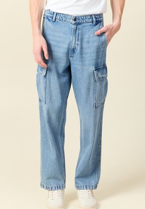 Pantalones cargo de mezclilla color azul claro con bolsillos laterales, un corte holgado y una textura ligeramente desgastada. Combinados con zapatillas blancas.