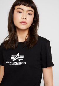 T-shirt en coton noir avec un logo graphique blanc bien en vue, des ailes et du texte, col rond et manches courtes.