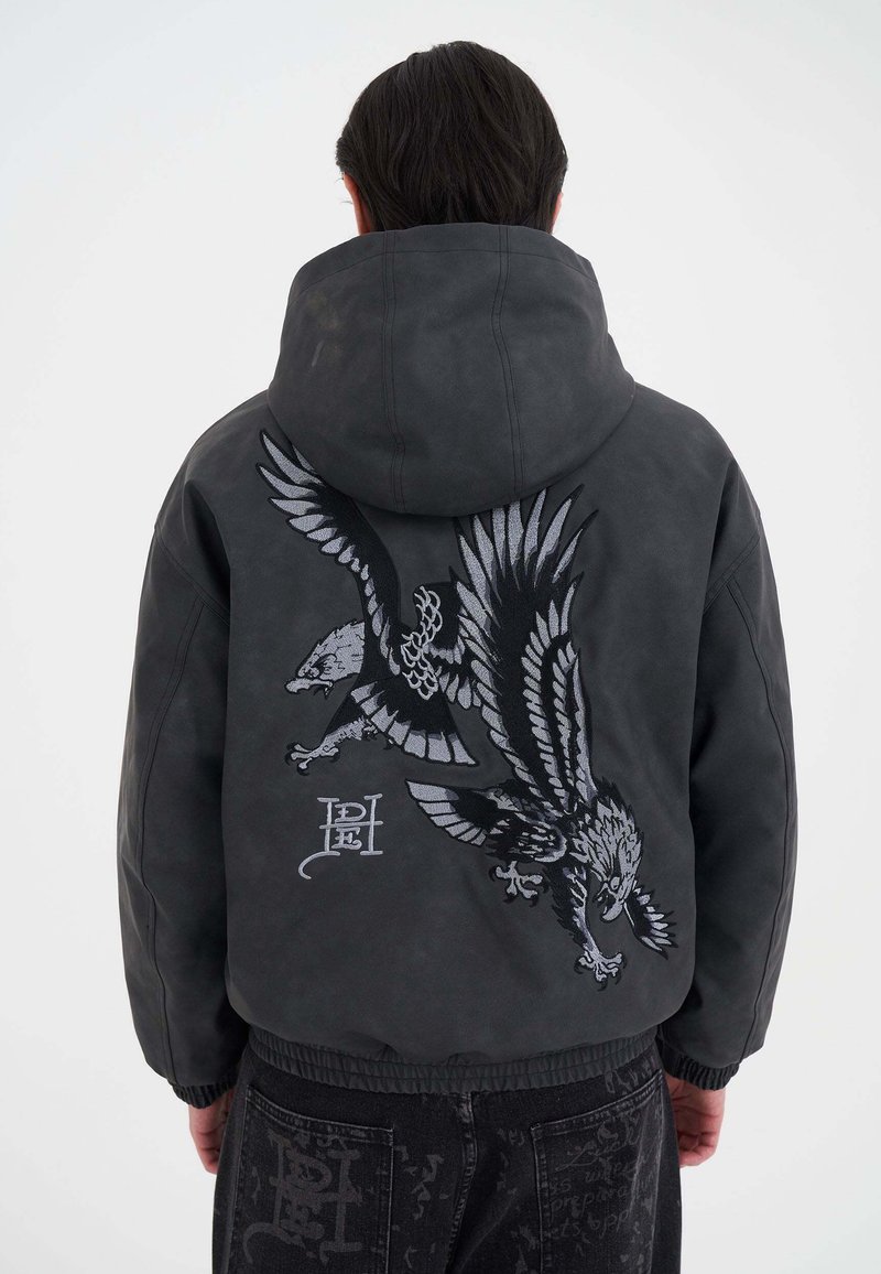 Persona che indossa una giacca con cappuccio grigio scuro con un grande design ricamato di due aquile in volo sulla schiena, abbinata a pantaloni neri con motivo.