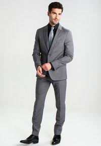 Abito grigio con vestibilità su misura, caratterizzato da una giacca monopetto, colletto a punta e pantaloni coordinati. Camicia nera, cravatta fantasia e scarpe lucide.