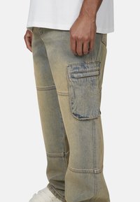 Mężczyzna w wyblakłych niebieskich dżinsach cargo z dużą kieszenią boczną, białych trampkach i białej koszuli, stojący na jednolitym tle.