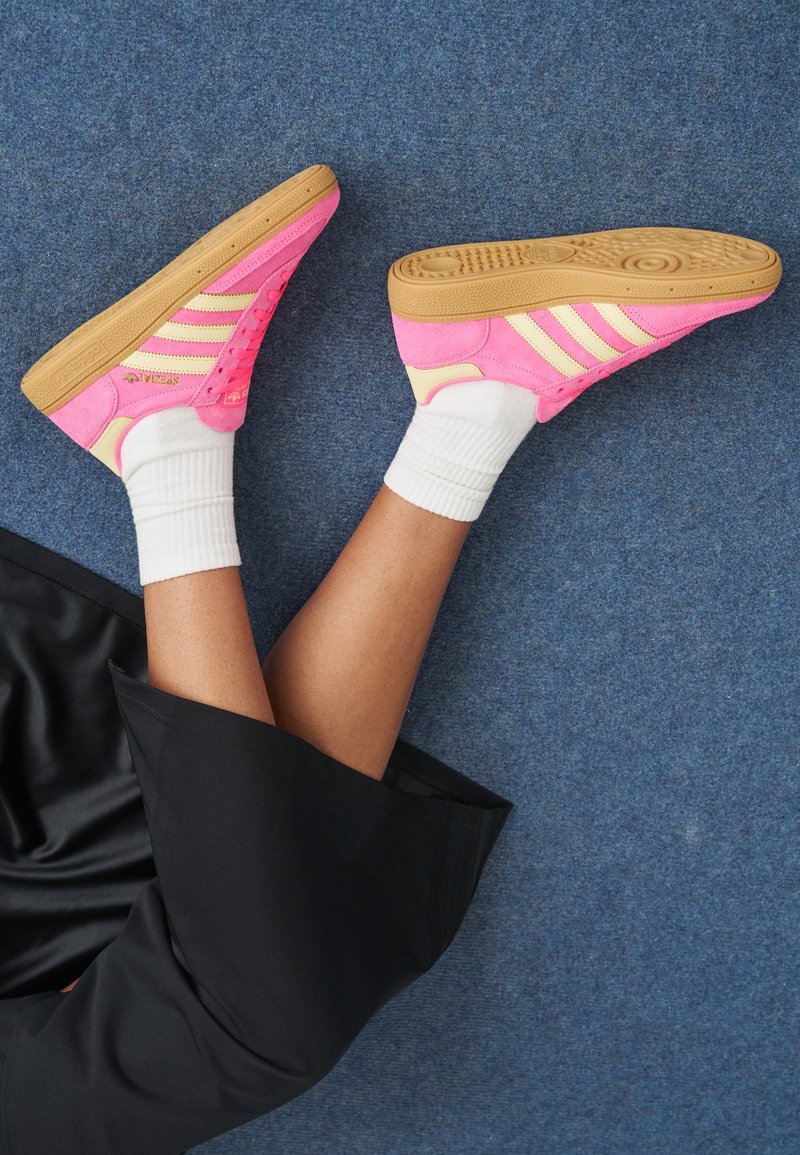 Person som bär rosa och beige Adidas-sneakers med vita strumpor, benen i kors, liggandes på en blå matta bredvid svart tyg.