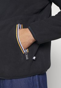 Giacca in pile nera con tasca con zip e accenti a strisce colorate. La giacca ha una finitura testurizzata e polsini a coste.