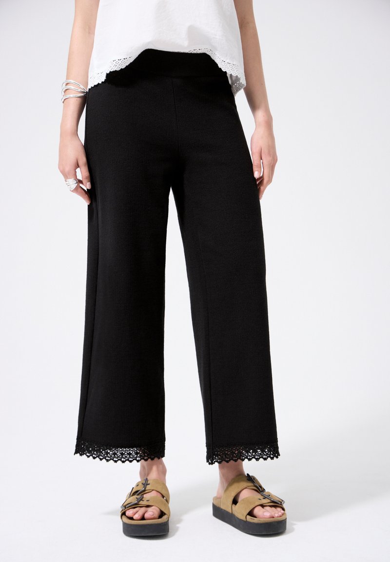 Femme portant un pantalon noir à jambes larges avec bordure en dentelle et des sandales compensées couleur naturel, debout devant un fond blanc.