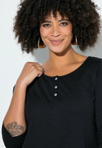 Haut noir à manches longues en matière douce, doté d'un décolleté en U et d'une patte à quatre boutons. Mis en valeur par un élégant tatouage sur le bras.