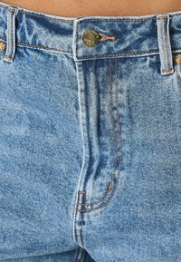 Primer plano de la cintura de unos jeans de mezclilla azul con un botón de latón etiquetado "ONLY" y costuras naranjas visibles.