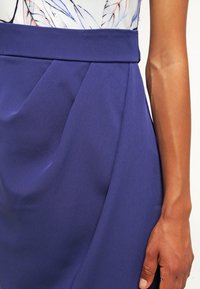 Milieu du corps d’une personne portant une jupe bleue taille haute plissée et un top blanc avec un motif de plumes, bras reposant le long du corps.