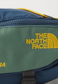 Borsa North Face con materiale verde testurizzato, logo e dettagli gialli, completata da una zip blu sulla parte superiore e hardware robusto.