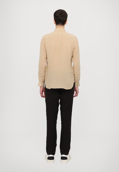 Camicia di lino beige a maniche lunghe con colletto, dettagli con bottoni, vestibilità su misura e orlo curvo. Indossata con pantaloni neri e sneaker casual.