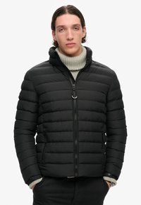 Sort polstret jakke med lynlås og høj krave; har et vandret quiltet design og sidelommer. Bæret over en lysegrå turtle neck.