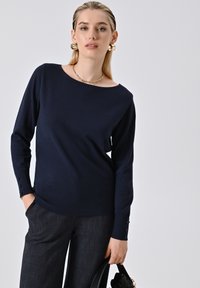 Marineblaues langärmeliges Sweatshirt mit weitem Ausschnitt und Knopfdetails an den Ärmeln, kombiniert mit dunkel strukturierten Hosen und einer schwarzen Handtasche.