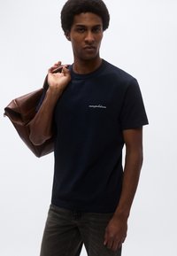 Tengerészkék pamut póló rövid ujjakkal és kerek nyakkivágással, fehér hímzett felirattal. A modell barna bőrtáskát tart. Látható farmernadrág.