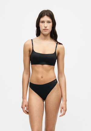 Femme debout face à l'avant portant un ensemble soutien-gorge et culotte noir en deux pièces avec un motif subtil et des bretelles de marque sur fond uni.
