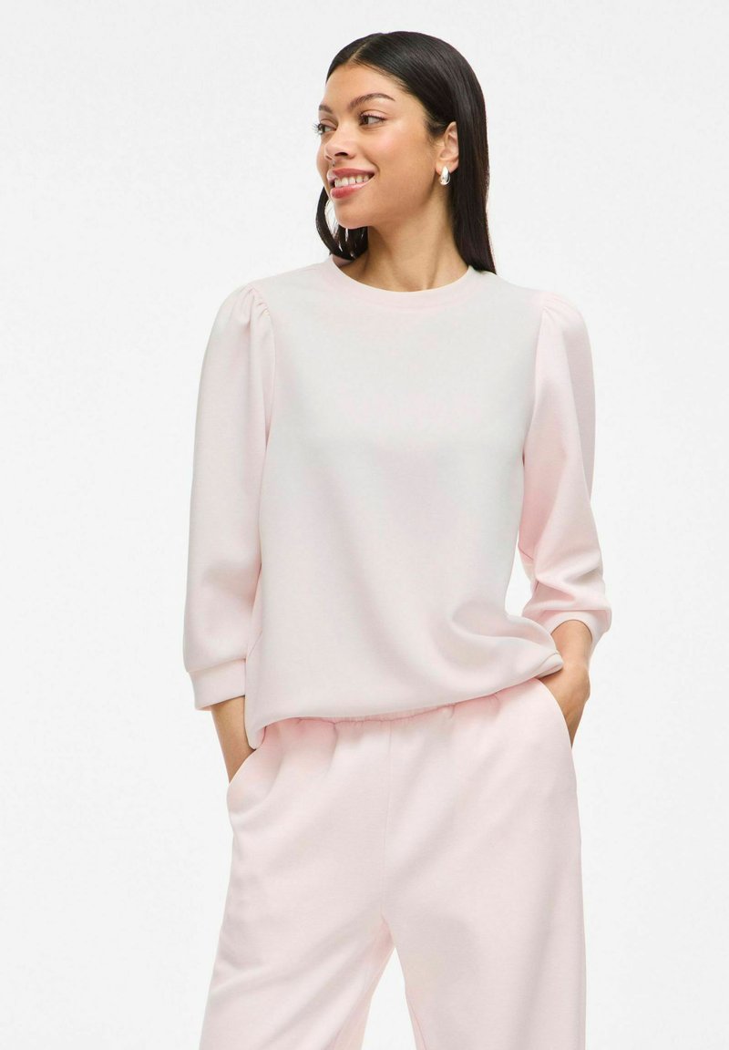 Blusa rosa chiaro a maniche lunghe con scollo tondo e spalle leggermente a sbuffo. Tessuto morbido con vestibilità rilassata, abbinata a pantaloni coordinati.