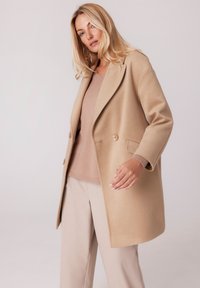 Beżowy płaszcz z mieszanki wełny z klapami, dwoma guzikami i bocznymi kieszeniami. Połączony z jasnobeżowym swetrem i dopasowanymi spodniami.