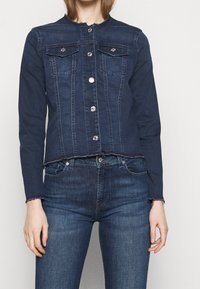 Chaqueta de mezclilla azul oscura con un diseño cortado, dos bolsillos en el pecho, botones a presión plateados y detalles deshilachados en el escote y en los puños.